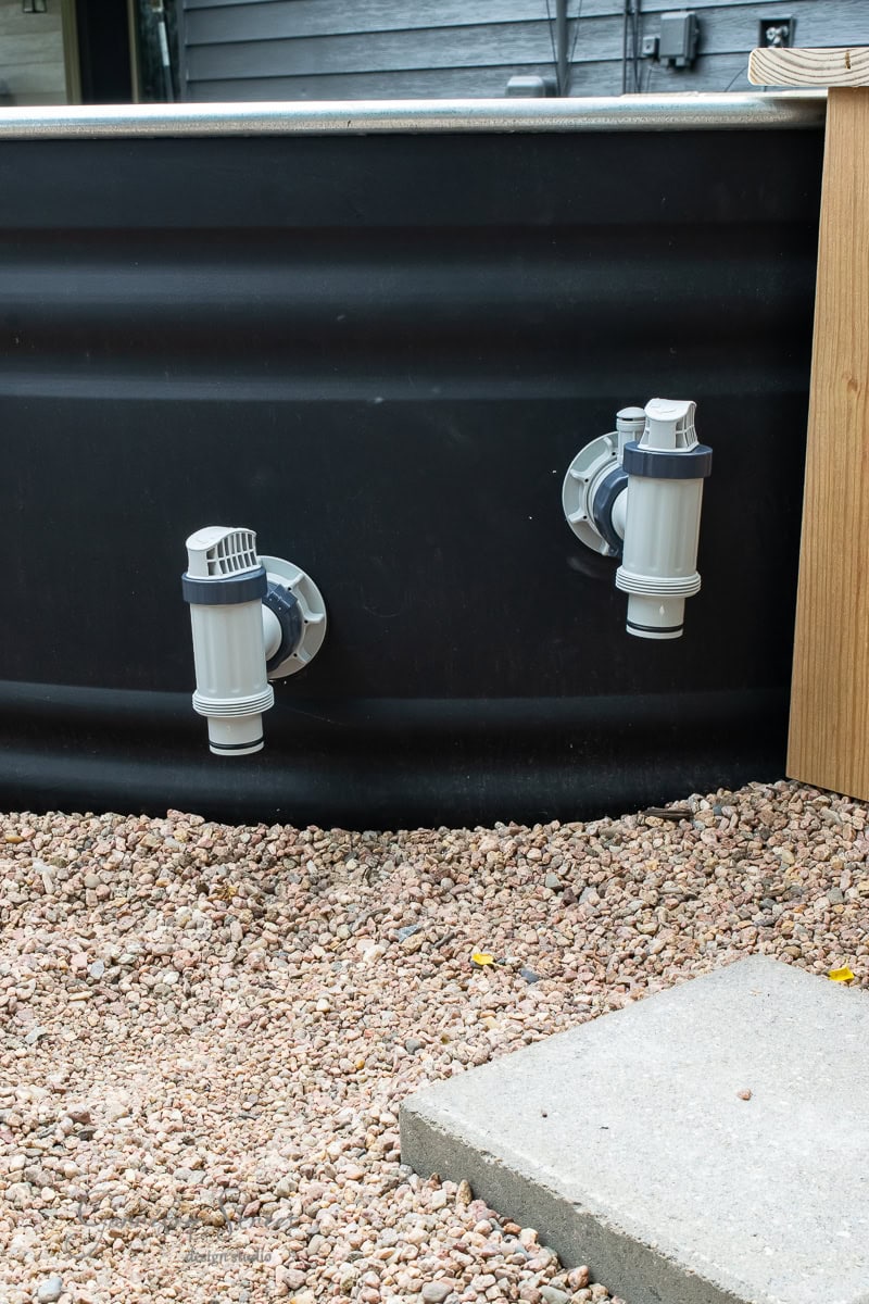 A close-up of two pool filter inlets attached to the side of a black above-ground stock tank pool. The ground around the pool is covered with small pebbles, and a concrete slab is partially visible in the bottom right corner.