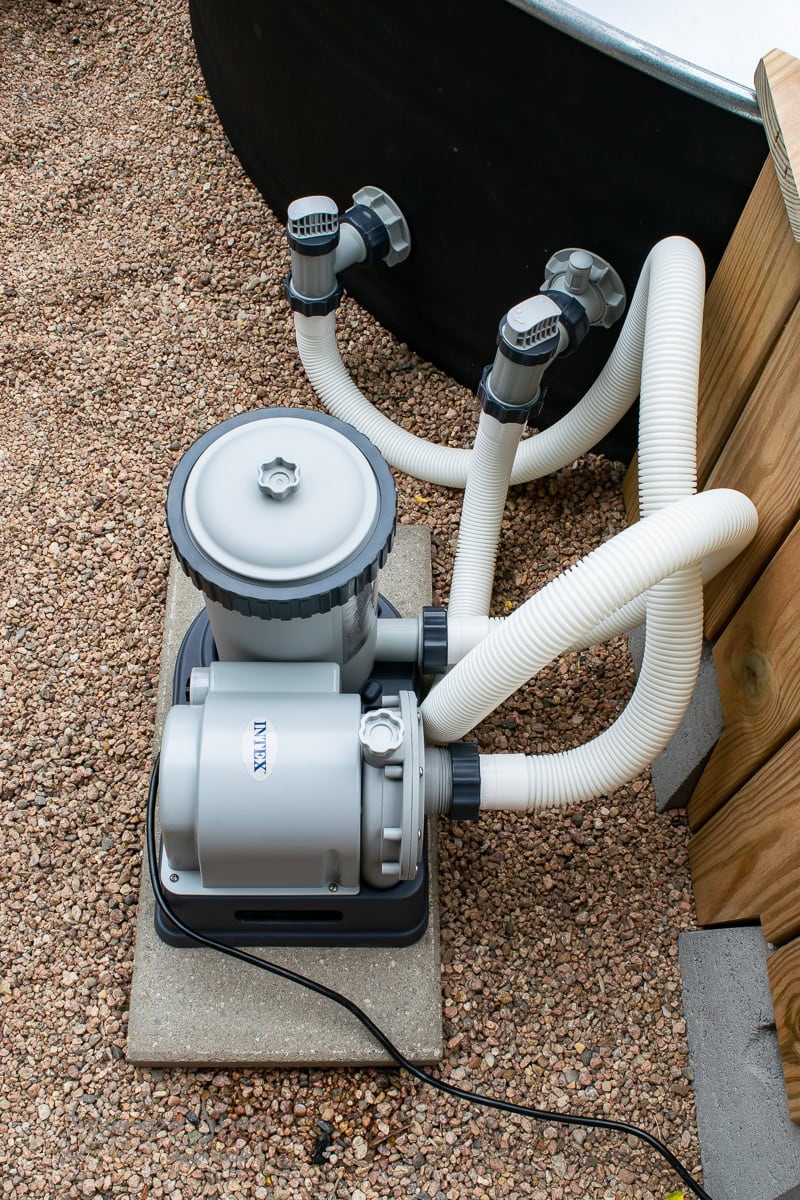 A gray pool filter system sits on a concrete slab, linked to a stock tank by white hoses. The setup is positioned on a gravel surface next to a wooden structure and a black container.