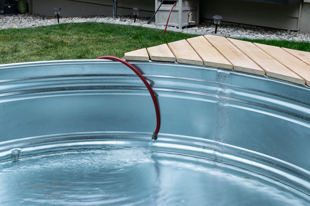 A stock tank pool is being filled with water from a red hose. It rests on the grassy lawn, snuggled up to a wooden deck in a charming backyard setting. The clear water sparkles invitingly under the sun.