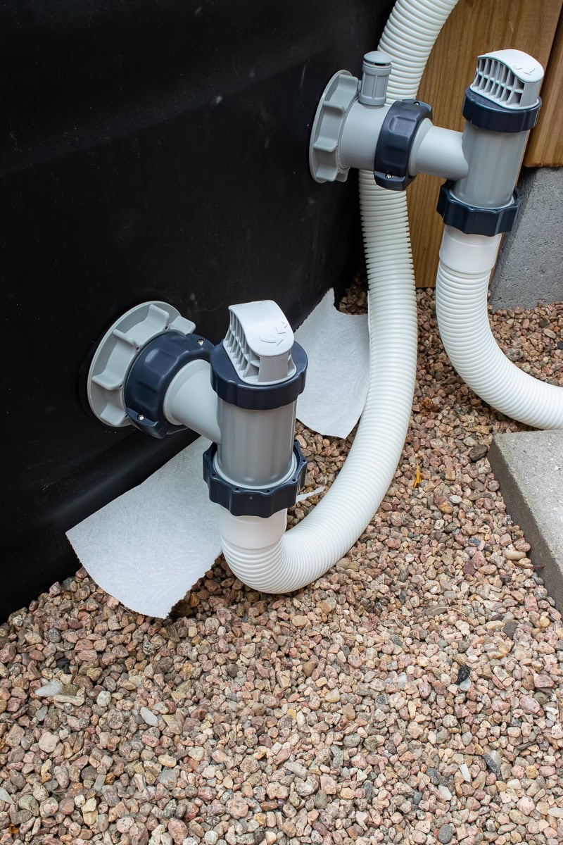 Close-up of a swimming pool pump and filtration system with hoses connected to a stock tank. The setup is placed on a bed of small pebbles, secured with paper padding underneath to stabilize the hoses.