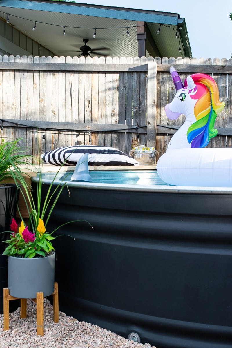 An inflatable unicorn float sits in a black stock tank pool in a fenced backyard. Nearby, a striped pillow rests on the pool's edge. A potted plant with colorful flowers is in the foreground, and string lights are hanging from the fence.