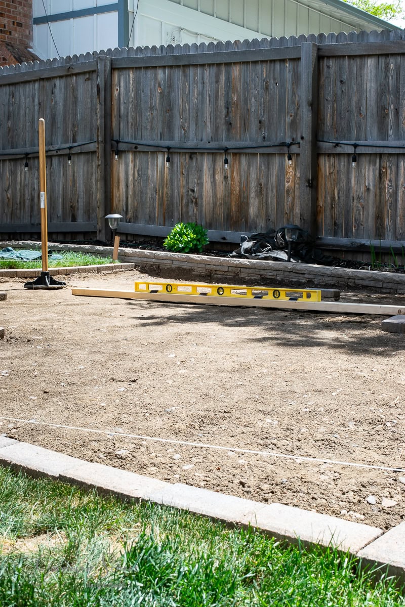 A backyard with a wooden fence and a garden bed in the background features a level on the ground and a tamper tool standing upright. In the foreground, grass lines the edge of the dirt surface, perfectly prepared for installing a stock tank pool.