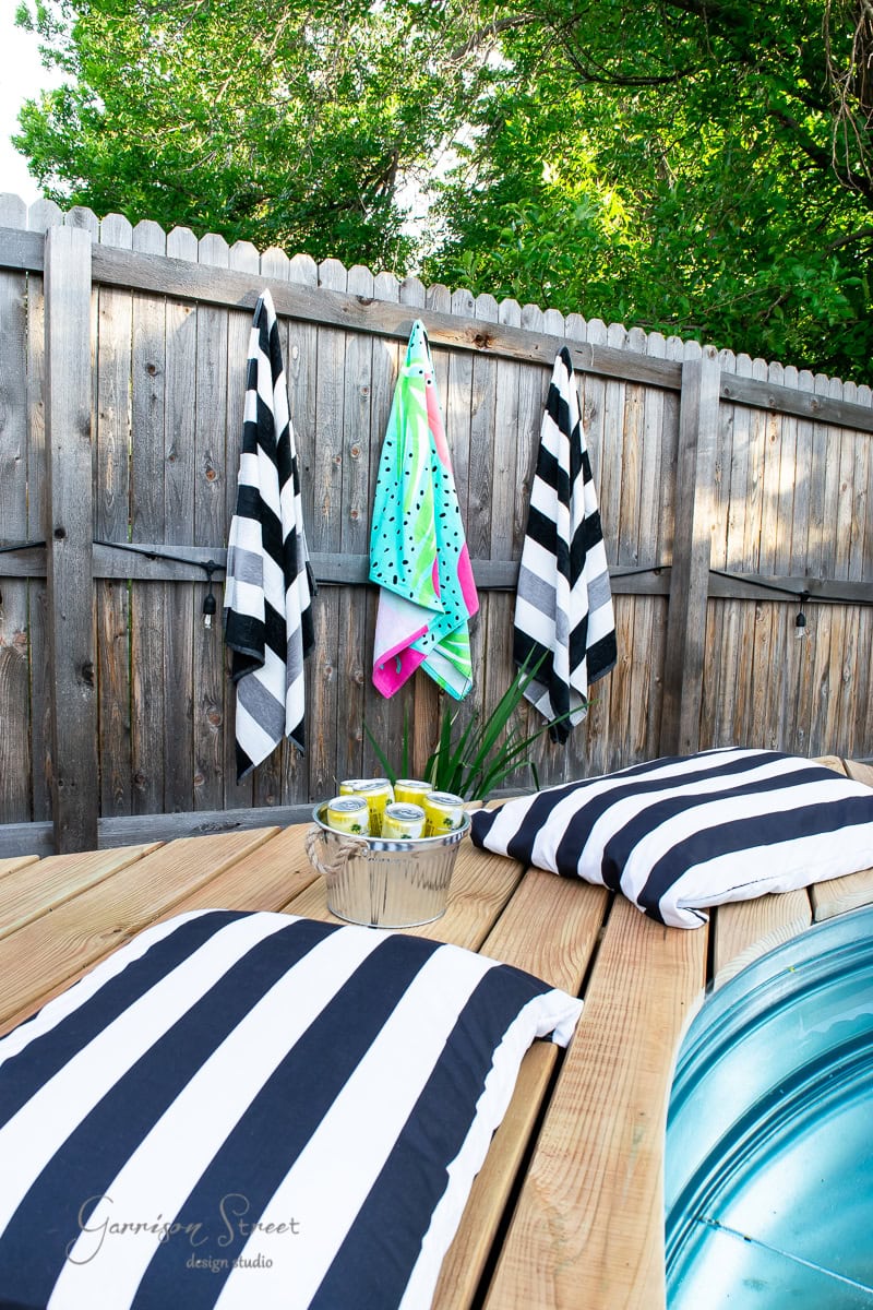 Outdoor picnic setup with black and white striped cushions on a wooden table, right next to a shimmering stock tank pool. Three striped towels and a colorful towel hang on a wooden fence. A metal bowl with beverages sits on the table, enveloped by lush greenery.