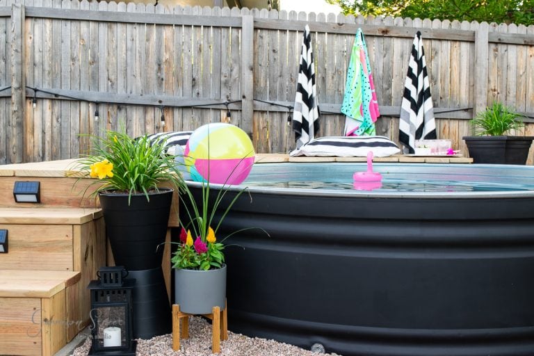 A small above-ground stock tank pool with colorful floats and striped towels on wooden chairs. Potted plants and a lantern decorate the area, with a wooden fence in the background. Steps lead up to the pool, surrounded by gravel.