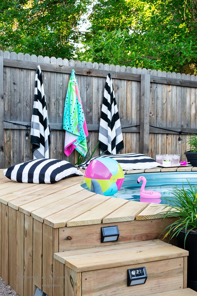 A small round stock tank pool is surrounded by wooden decking. Black and white striped towels and cushions are placed around. A colorful beach ball, a pink flamingo float, and green plants add to the vibrant setup. A wooden fence and foliage are in the background.