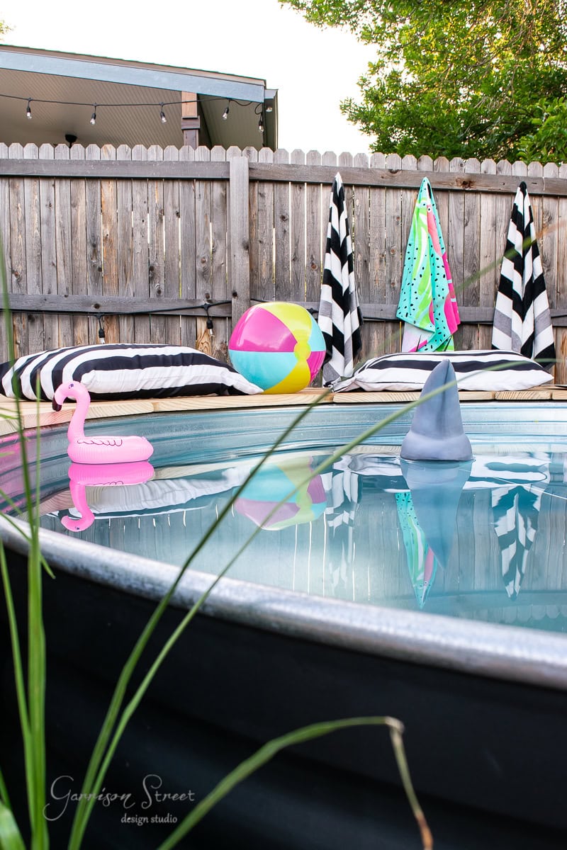 A small backyard pool with clear water features an inflatable pink flamingo and a gray shark fin. Striped towels hang on the wooden fence, while colorful cushions and a beach ball rest by the deck. A sense of serenity is enhanced by lush greenery visible in the background.