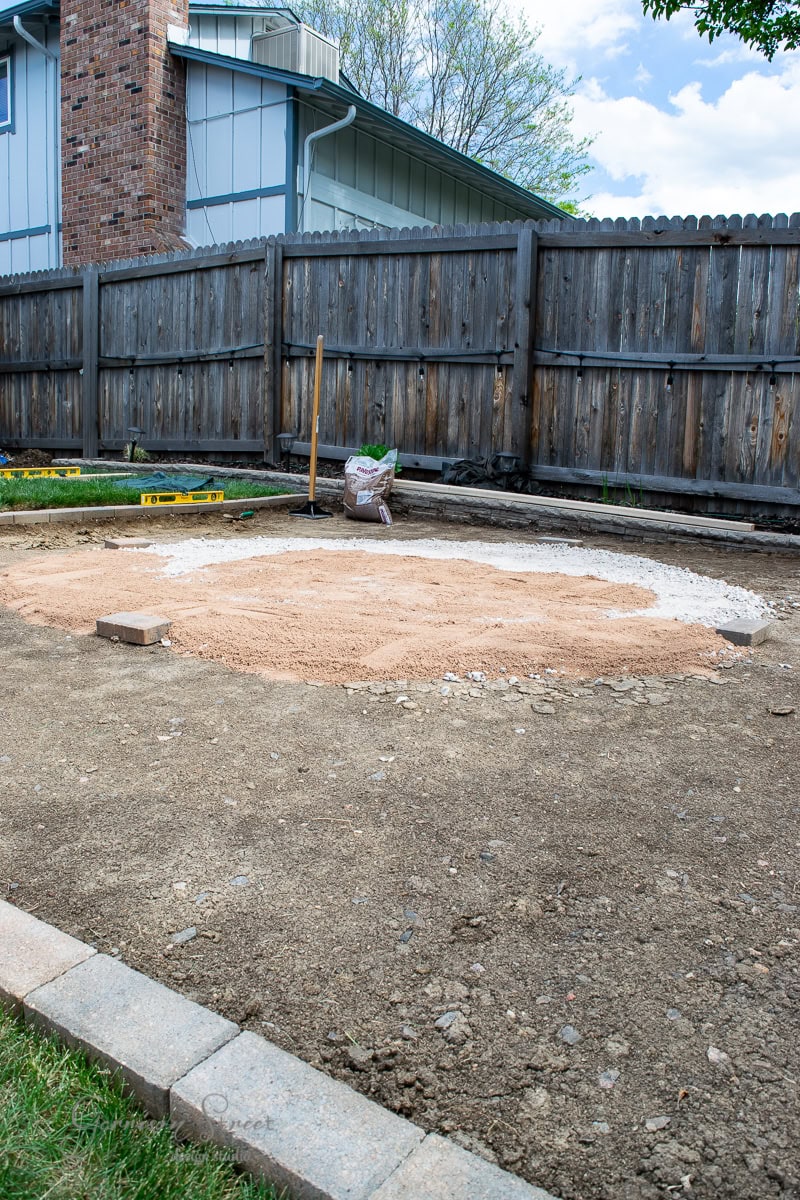 A backyard with a partially prepared round area of sand and gravel for a future pool or stock tank, surrounded by a wooden fence. Some building tools, including a shovel and a level, are visible. A patch of grass is in the lower left corner.