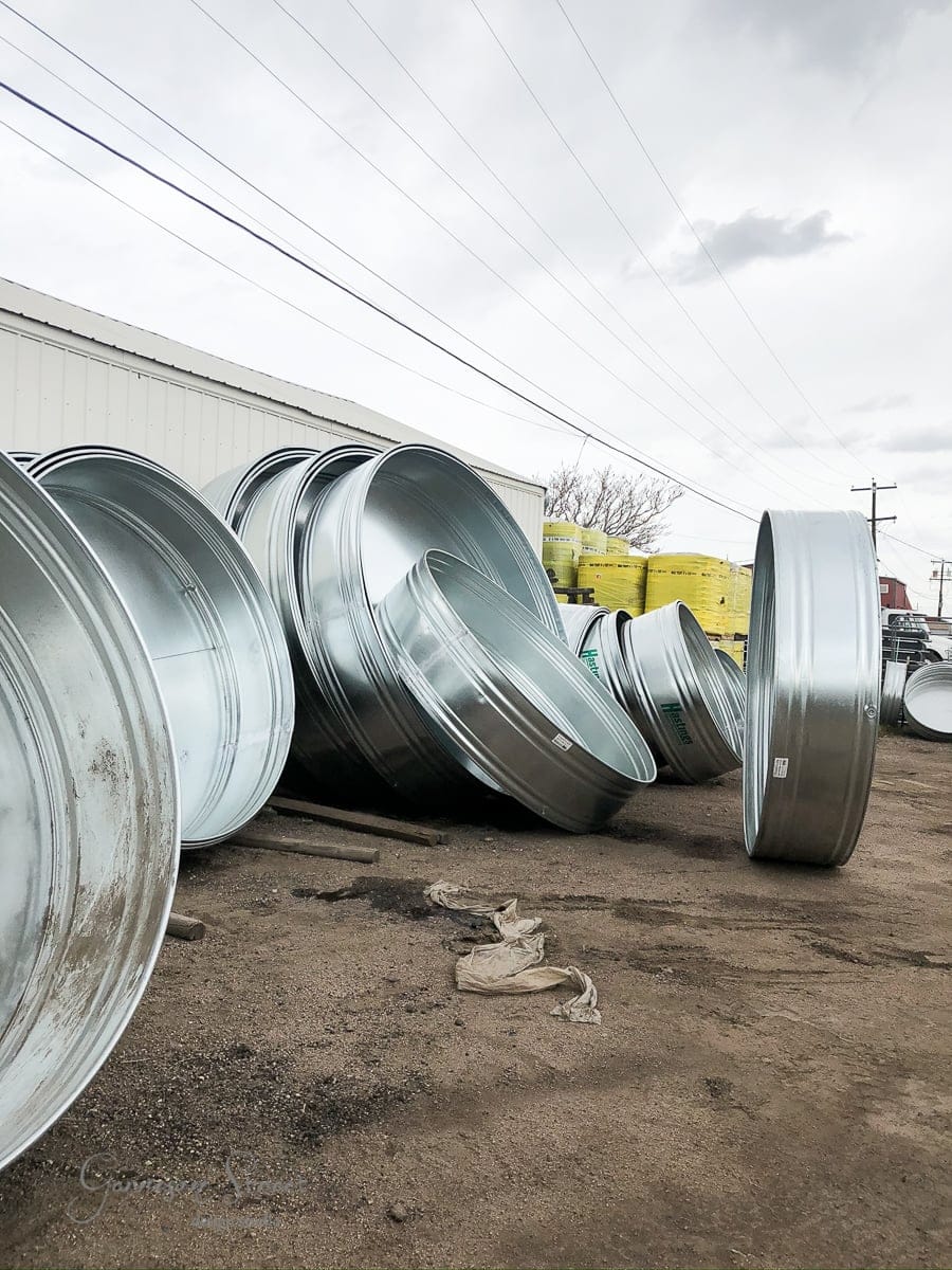 Stacks of large, round metallic containers, resembling oversized stock tanks, are scattered on a dirt ground outside a warehouse. Power lines and an overcast sky loom in the background, lending an industrial feel to the scene.
