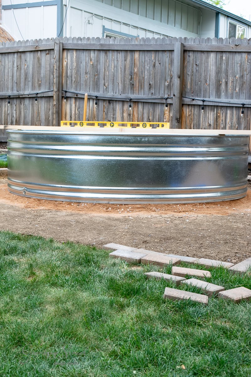 A large, silver stock tank pool sits on bare ground in a backyard, complemented by a wooden fence in the background. A yellow level tool rests atop the tank. Green grass and scattered bricks add charm to the foreground.