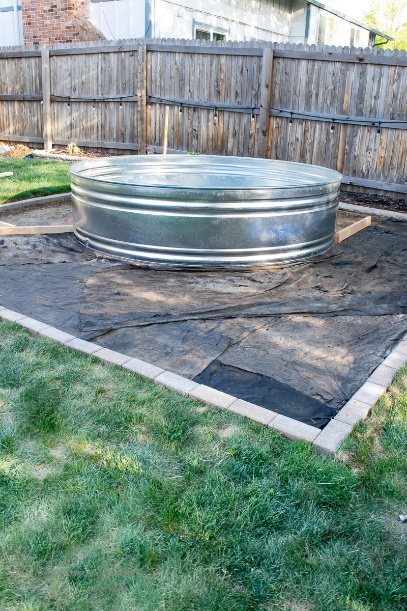 A large metal stock tank pool is set up in a backyard with a wooden fence. It's placed on a tarp-covered area framed by bricks. The surrounding ground is grassy, and the sky is clear.