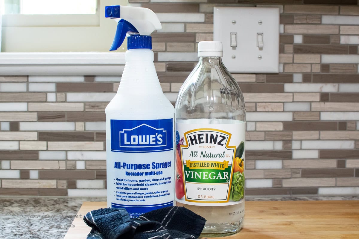 A blue spray bottle labeled "All-Purpose Sprayer" stands next to a clear bottle of Heinz distilled white vinegar on a wooden countertop, reminiscent of prepping a stock tank pool for summer. A dark cloth lies beside them, with a tiled backsplash in the background.