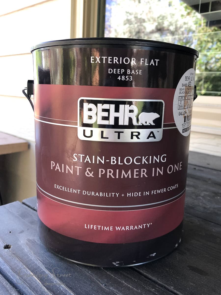 A gallon can of Behr Ultra exterior flat paint and primer in deep base sits on a wooden surface, perfect for projects like sprucing up a stock tank. The label highlights features such as stain-blocking, excellent durability, and lifetime warranty.