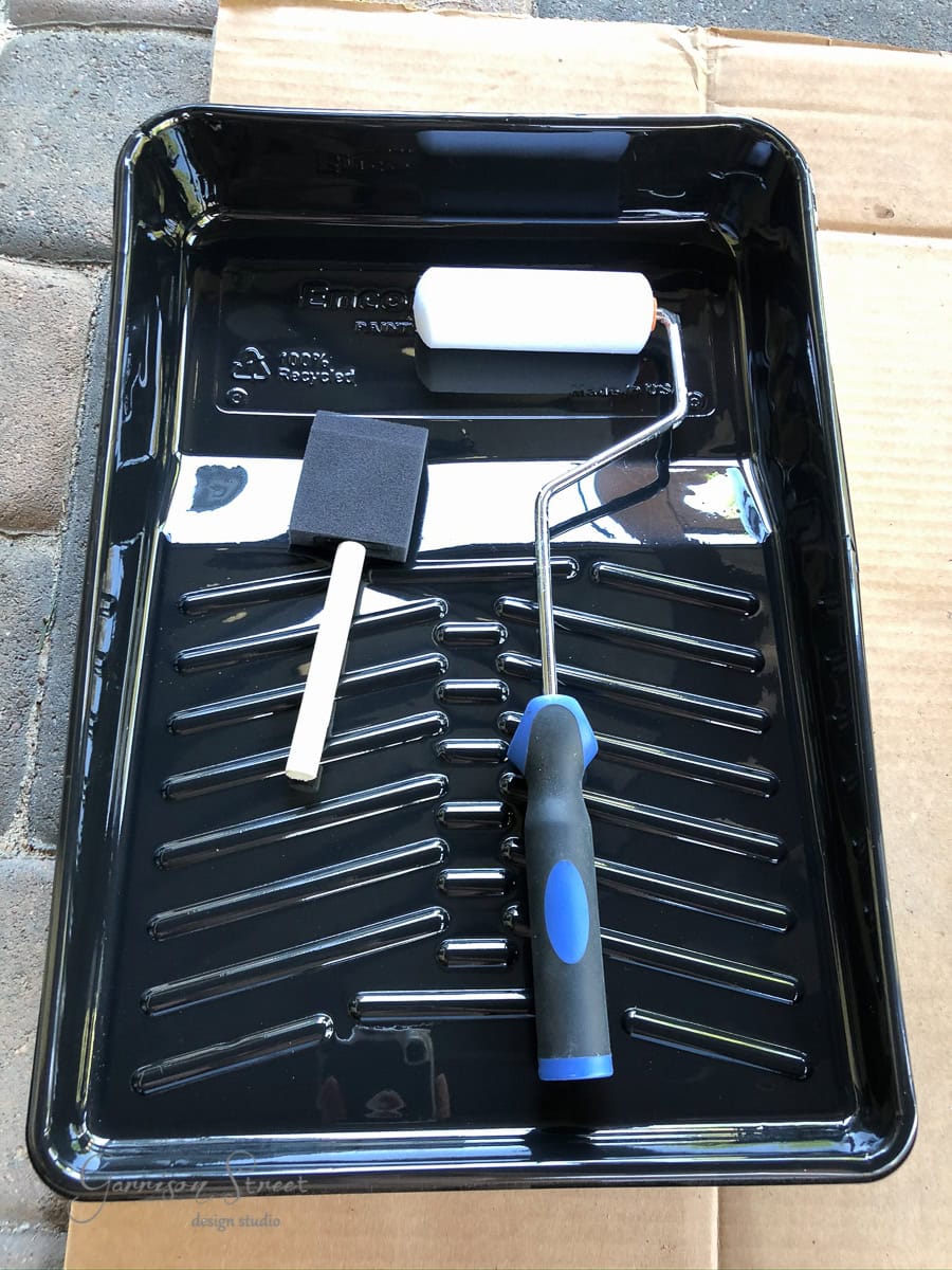 A black paint tray rests on cardboard, holding a foam paint roller with a black and blue handle, reminiscent of the vivid hues of a pool. Accompanying it is a black foam brush with a wooden handle, ready for any painting task.