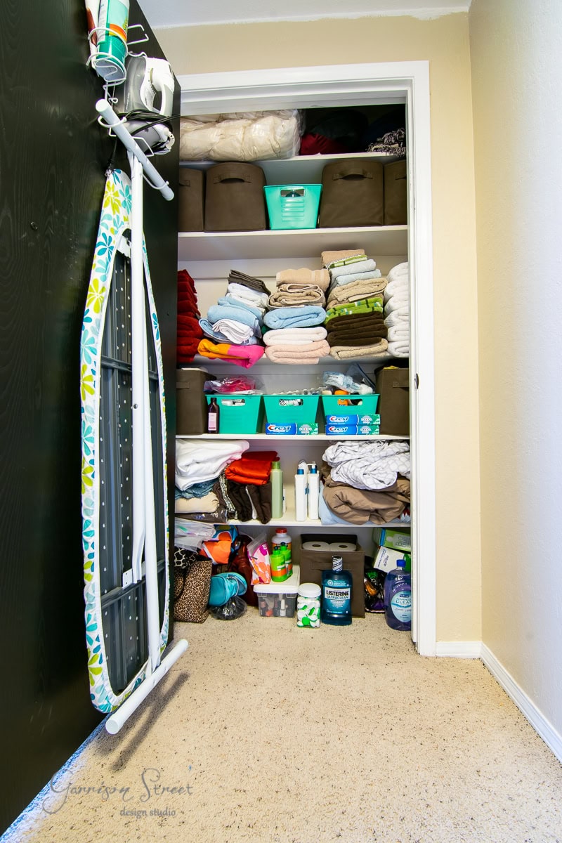 Shelves brimming with folded towels, bins, and cleaning supplies. An ironing board with a colorful cover hangs on the inside of the door. The carpeted floor complements the closet's light beige wall.