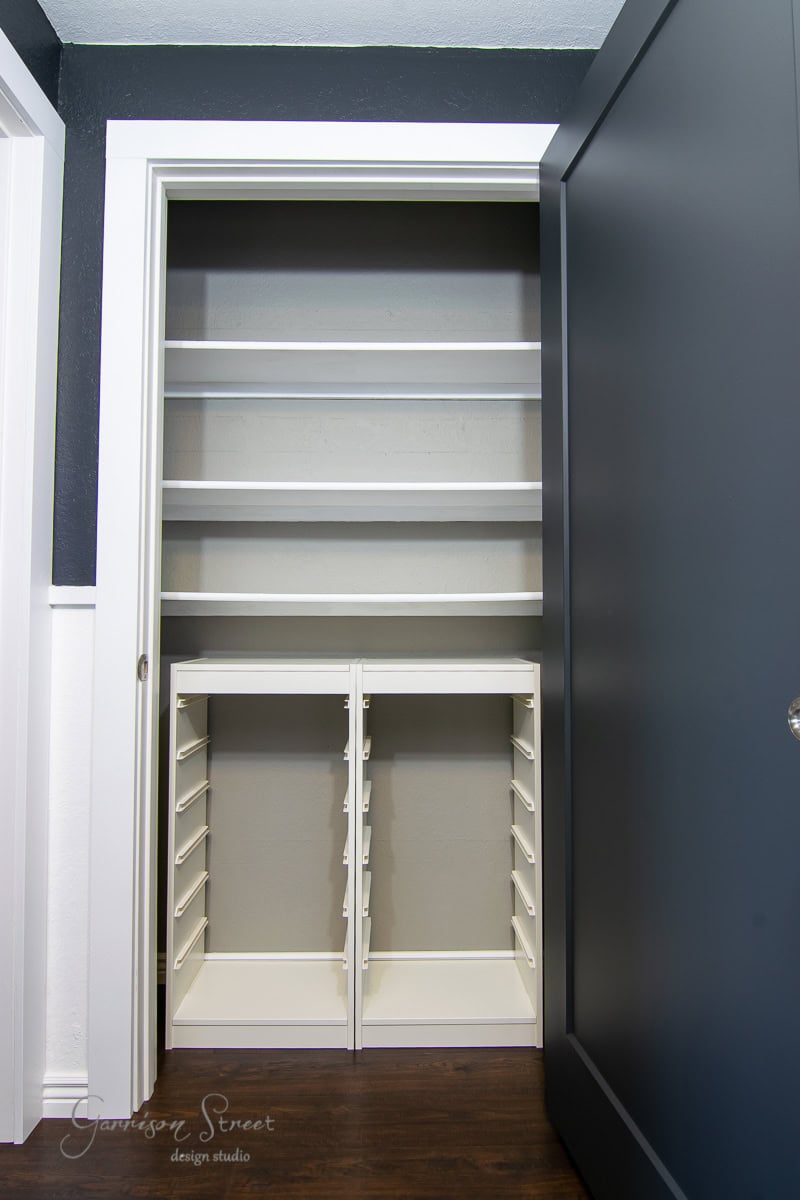 A small, open linen closet features an elegant makeover with white shelves on both sides. The left boasts four wide shelves, while the right is accented by a sleek black sliding door. The empty space is completed by a warm, wooden floor beneath.