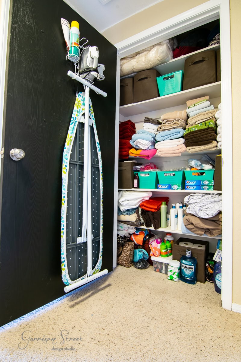 Shelving unit with towels and linens stacked on the shelves, alongside plastic bins for storage. An ironing board hangs on the inside of the door, while various cleaning supplies rest on the floor.