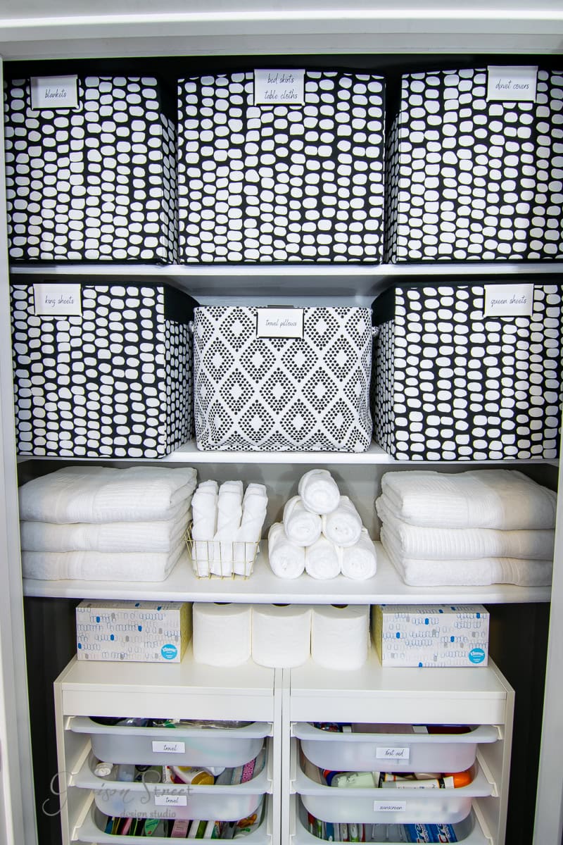 Neatly organized shelves featuring labeled black and white patterned storage bins. Towels are stacked meticulously, and tissues rest in elegant boxes. Drawers hold a spectrum of washcloths, all arranged with precision.