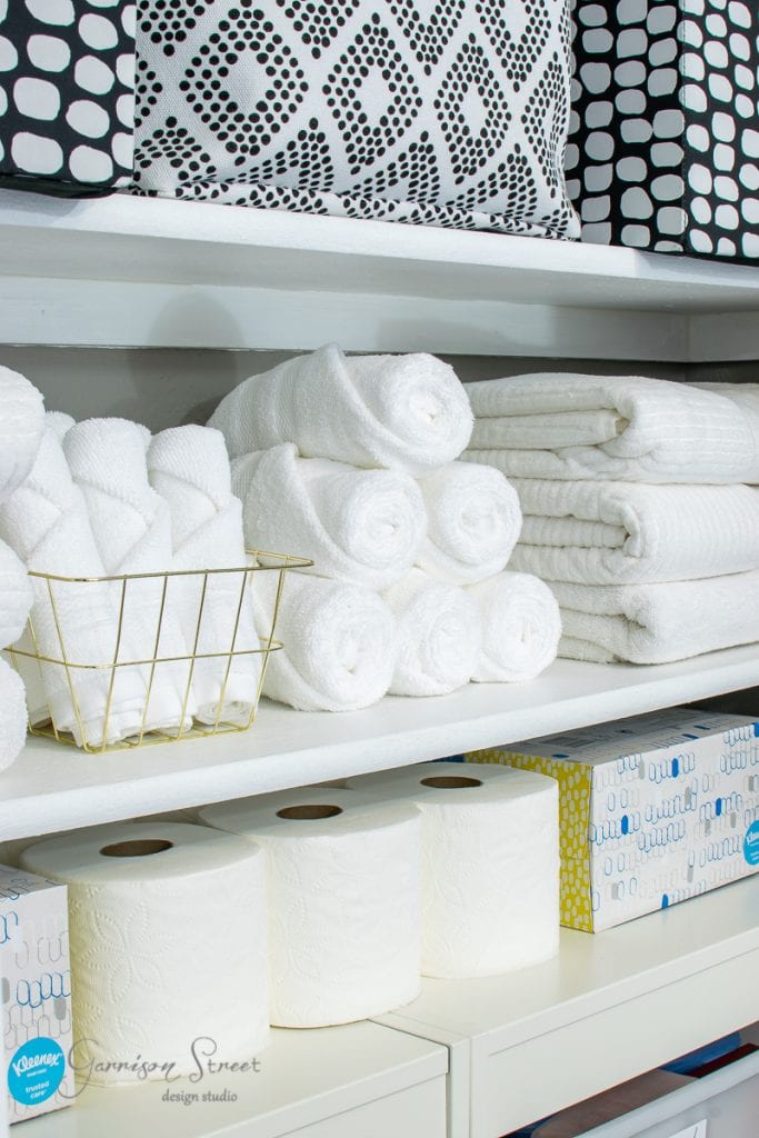 Home Shelves neatly stocked with rolled white towels, a basket of hand towels, toilet paper rolls, tissue boxes, and black-and-white patterned storage bins on the top shelf showcase the results of a stunning linen closet makeover.