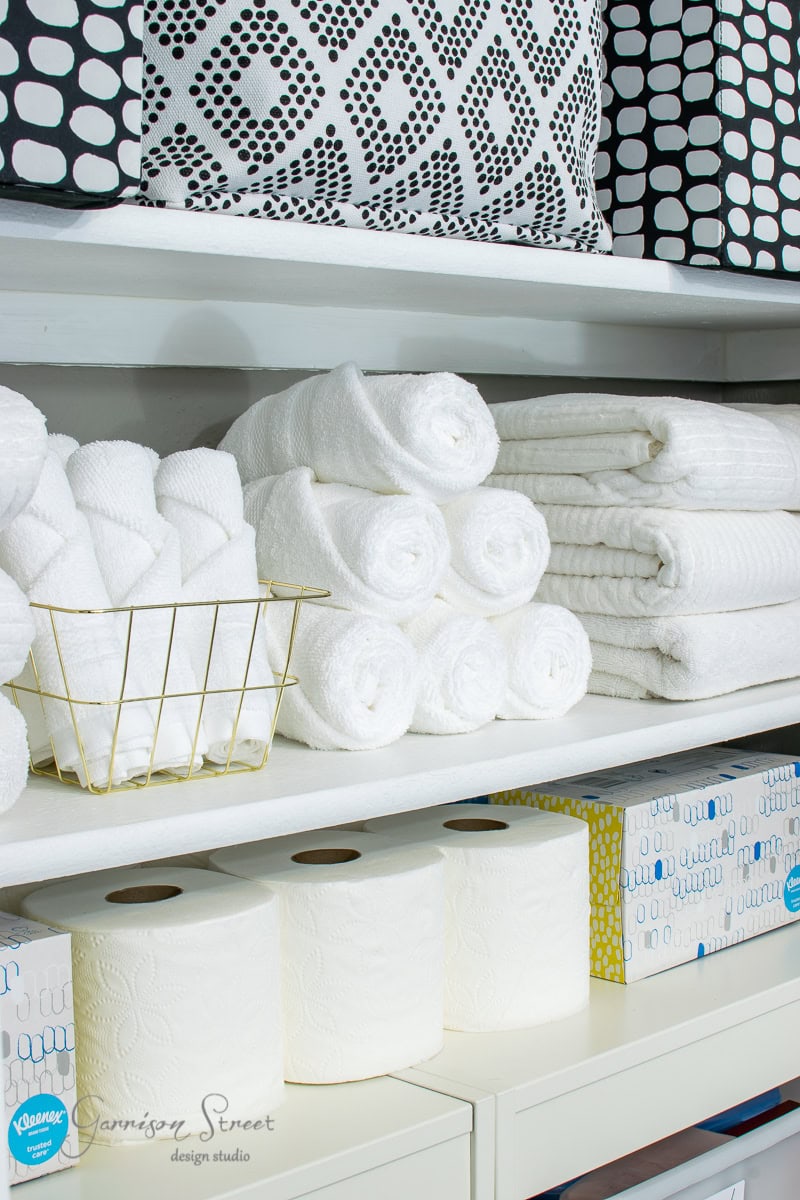 Shelves neatly stocked with rolled white towels, a basket of hand towels, toilet paper rolls, tissue boxes, and black-and-white patterned storage bins on the top shelf.