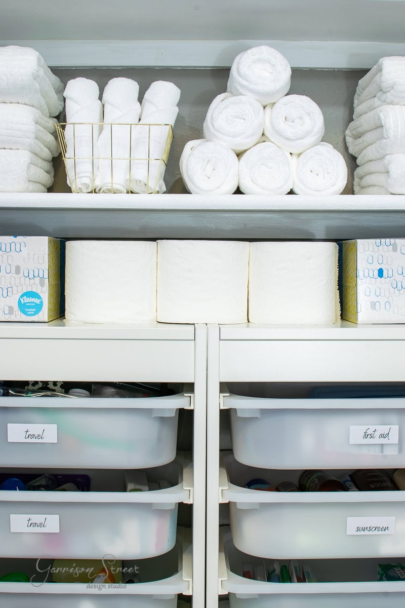 Rolled and stacked white towels on top. Below, a row of toilet paper sits above white drawers labeled for travel items, first aid, and sunscreen. Everything is orderly and accessible.