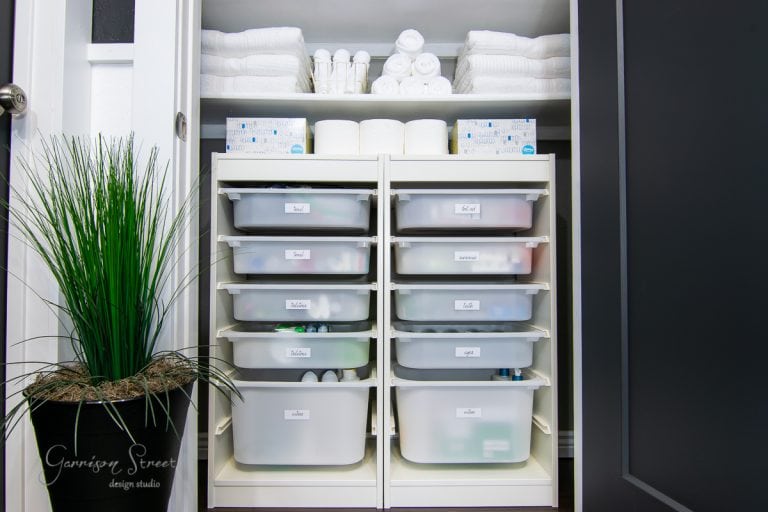 A neatly organized linen closet makeover features two shelving units with pull-out bins labeled for storage. The top shelf holds stacks of white towels, rolls of toilet paper, and toiletries. A potted plant is placed to the left of the open closet.