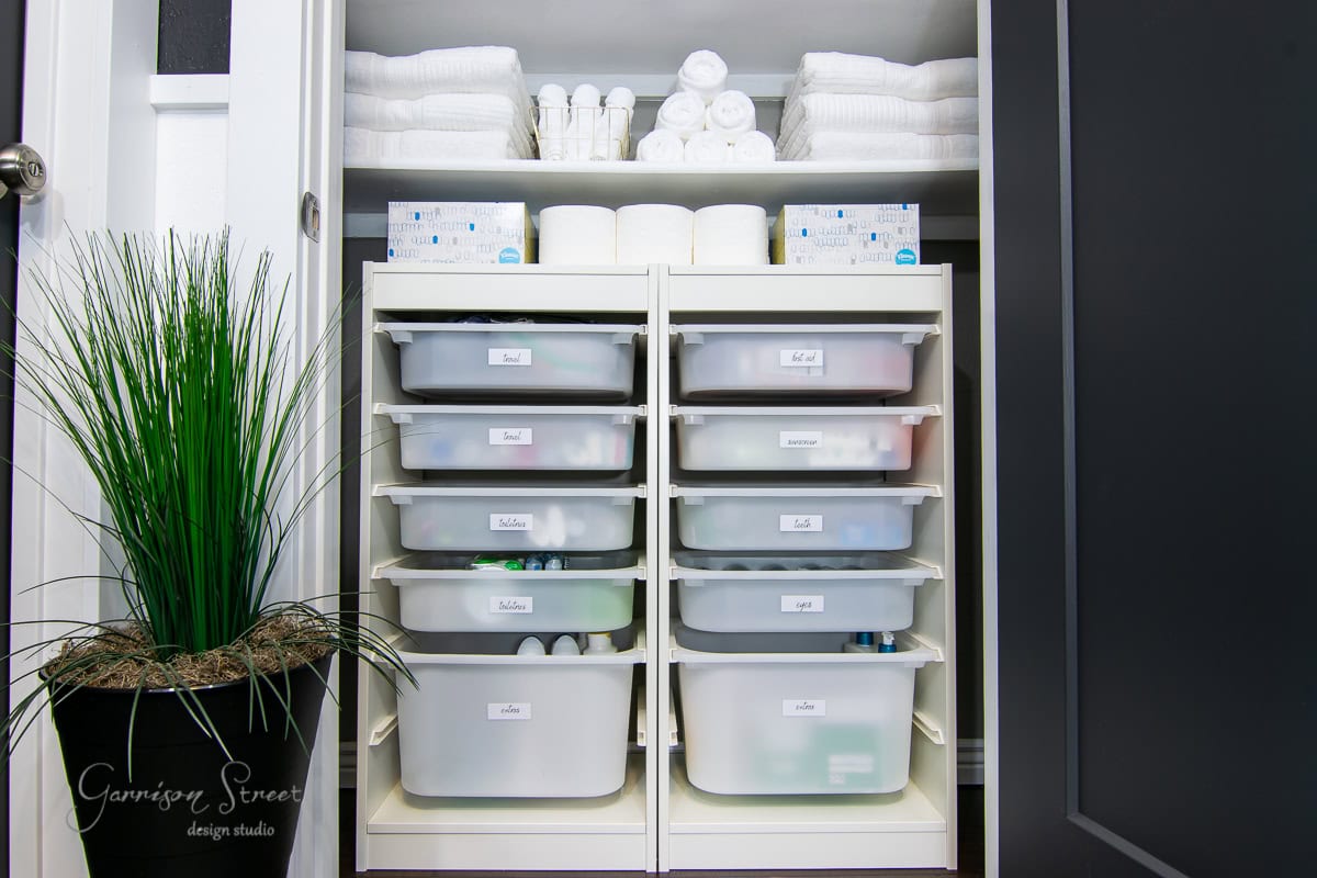 A neatly organized linen closet makeover features two shelving units with pull-out bins labeled for storage. The top shelf holds stacks of white towels, rolls of toilet paper, and toiletries. A potted plant is placed to the left of the open closet.