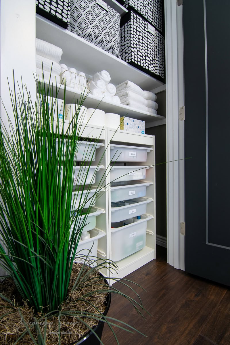 Shelves of neatly stacked towels, chic black and white patterned baskets, and labeled plastic drawers. A green potted plant adds a fresh touch against the rich dark wooden floor.