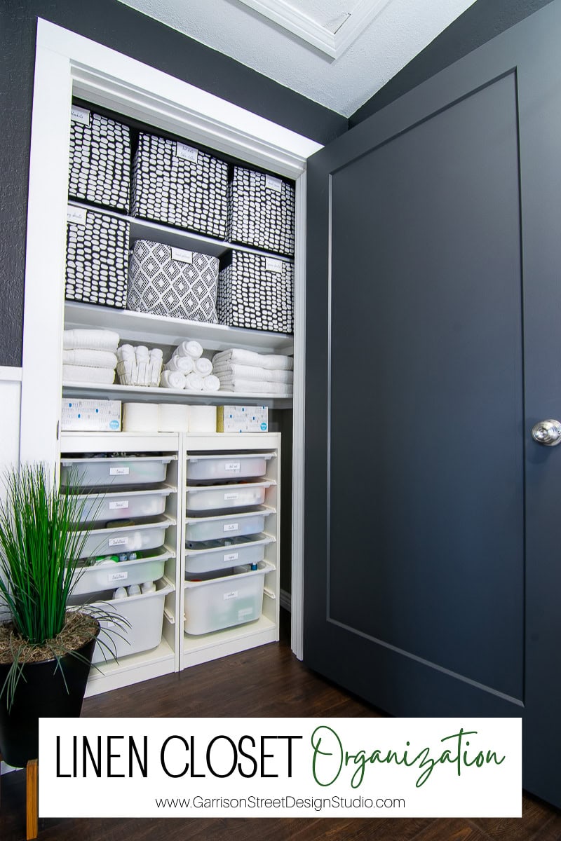 A neatly organized linen closet makeover showcases stacked gray and white patterned towels on top shelves. The middle shelves hold folded white towels and rolls, while labeled storage bins sit below. A small plant adds a touch of greenery beside the open closet door.