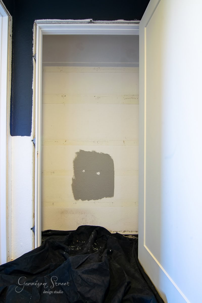 An empty linen closet with partially painted walls showcases a gray paint swatch over an off-white backdrop. The door stands open, hinting at a makeover in progress, while a black drop cloth neatly covers the floor.