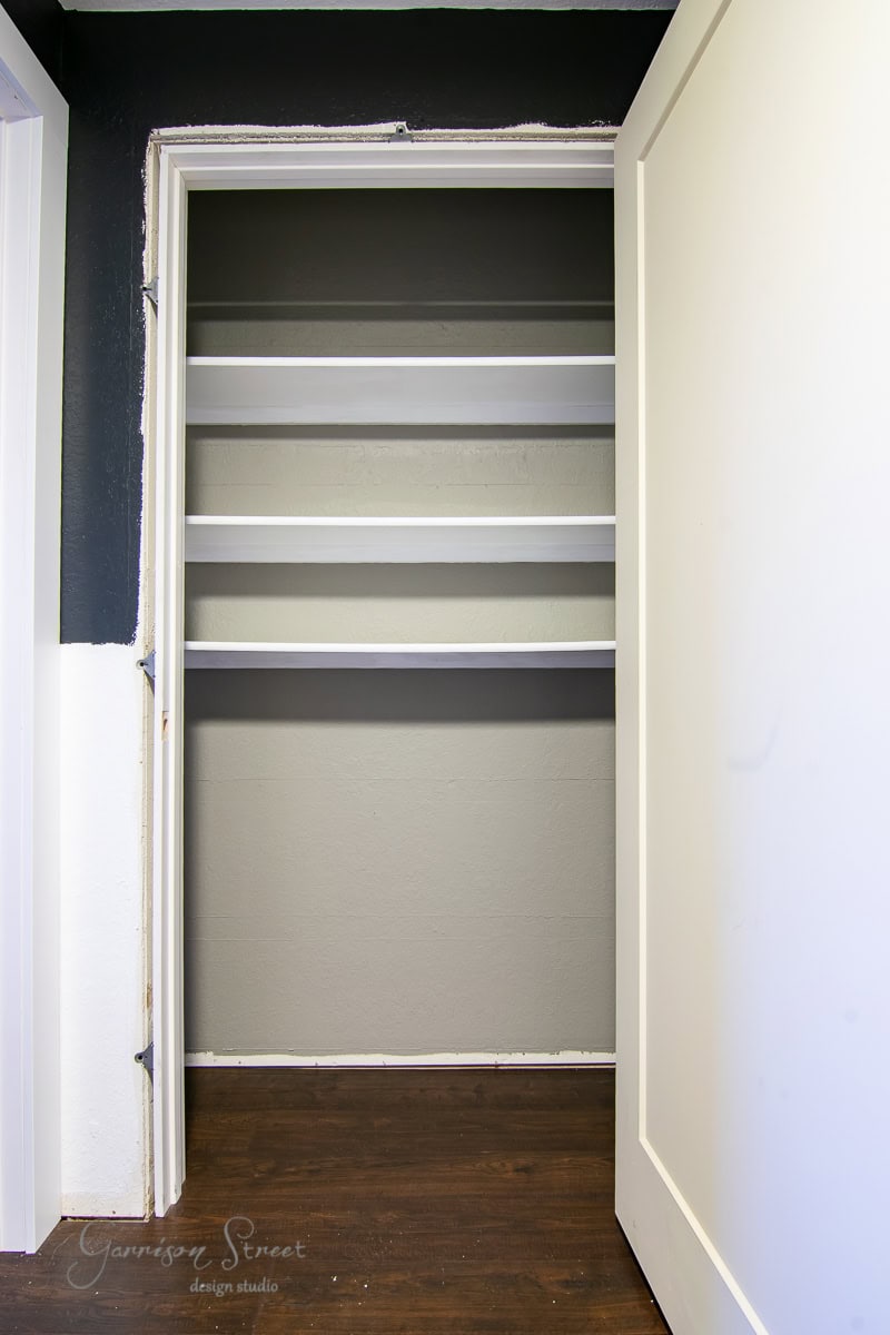 An empty closet with four white shelves stands against a two-tone wall with dark upper and light lower sections. The white door complements the space as it rests on a dark hardwood floor.