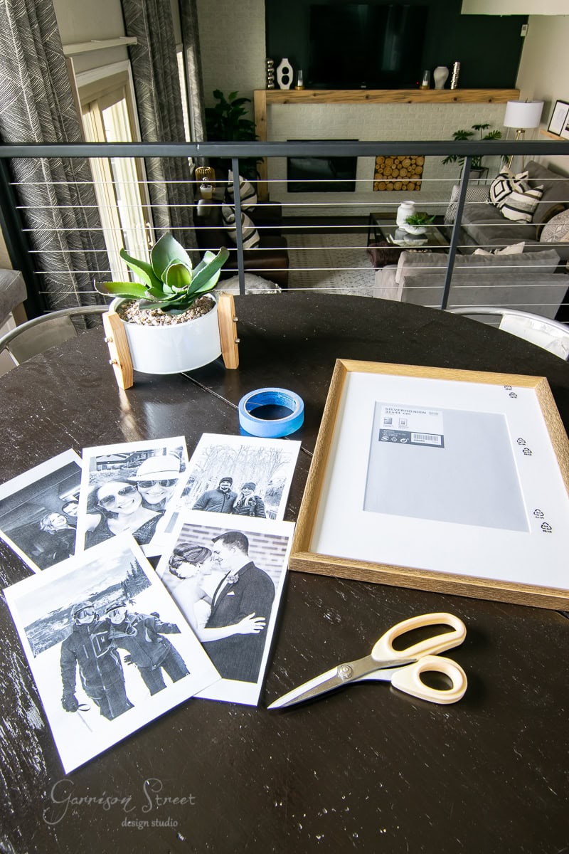 A collection of black and white photos spread on a dark round table alongside a pair of scissors, blue tape, an oversized picture frame, and a potted plant. A modern living room with a TV and fireplace is in the background.