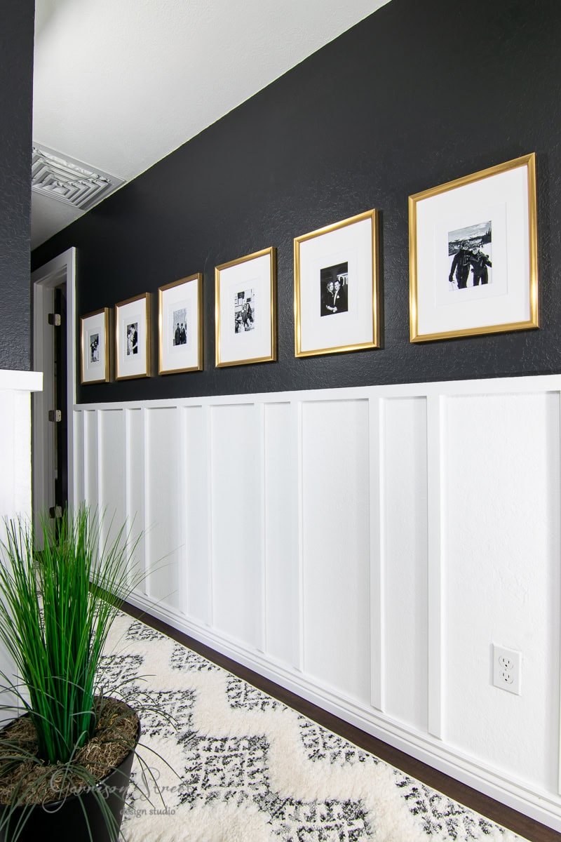 A hallway with black walls and white wainscoting features six framed black-and-white photos in oversized gold picture frames. A green plant is positioned near a patterned black-and-white mat on the floor.