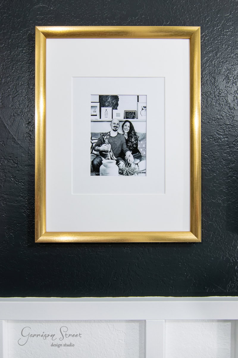 An oversized black and white photo hangs on a textured dark wall, encased in a golden picture frame with a delicate mat. The image captures two people sitting on a couch with their dog. Beneath the frame, "Garrison Street Design Studio" is subtly inscribed on the wall.
