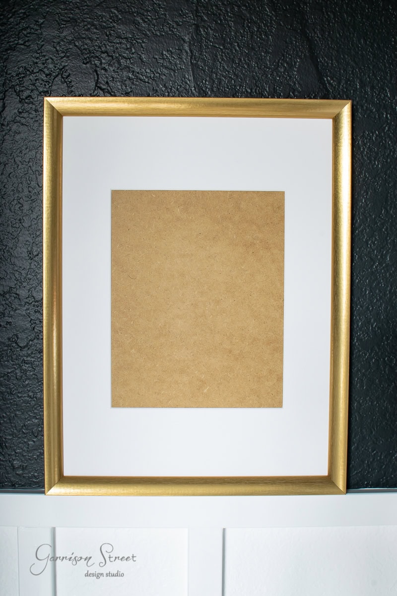 An oversized gold-framed picture frame hangs on a textured black wall. The frame houses a blank brown paper in the center, gracefully bordered by a white mat. In the lower left corner of the wall, a small signature reads "Garrison Street Design Studio."