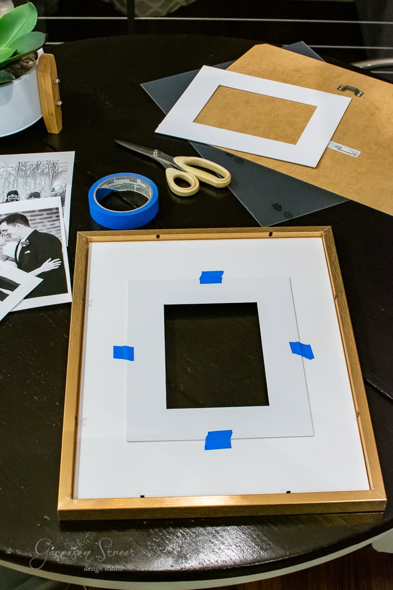 An oversized tabletop displays a picture frame, scissors, and blue painter's tape for mounting a photo. A black-and-white photo and an oversized mat board are visible, alongside additional supplies like black paper and an adhesive roll.