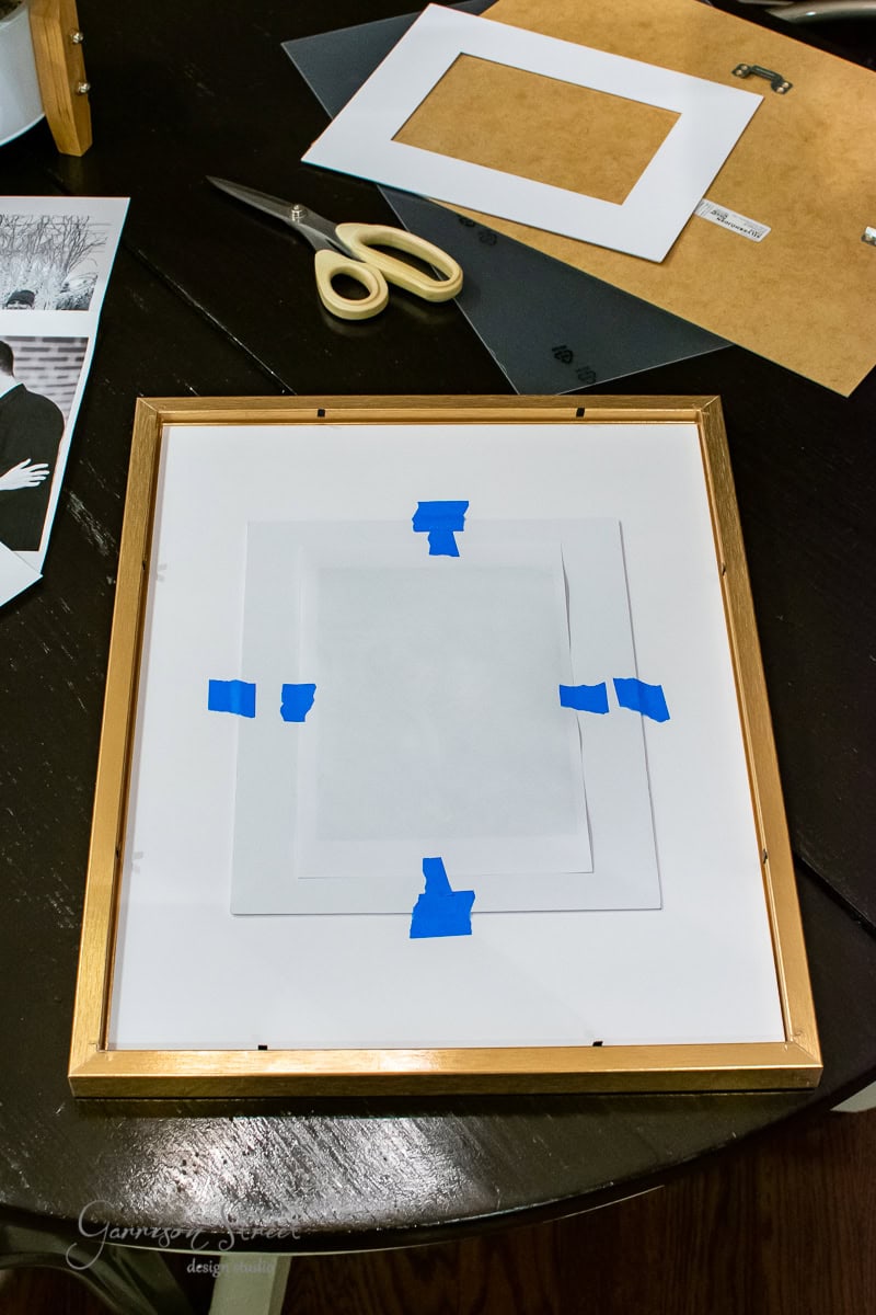 An oversized wooden picture frame lies on a table, with a photo mat neatly taped in the center. Scissors, a paper cutter, and an envelope are nearby. Partially visible black and white photos scatter across the surface.