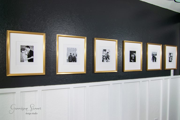 A series of six black and white photos in gold picture frames, accented with oversized mats, are neatly arranged on a black wall with white wainscoting. Each frame contains a different portrait, creating a cohesive, elegant display.
