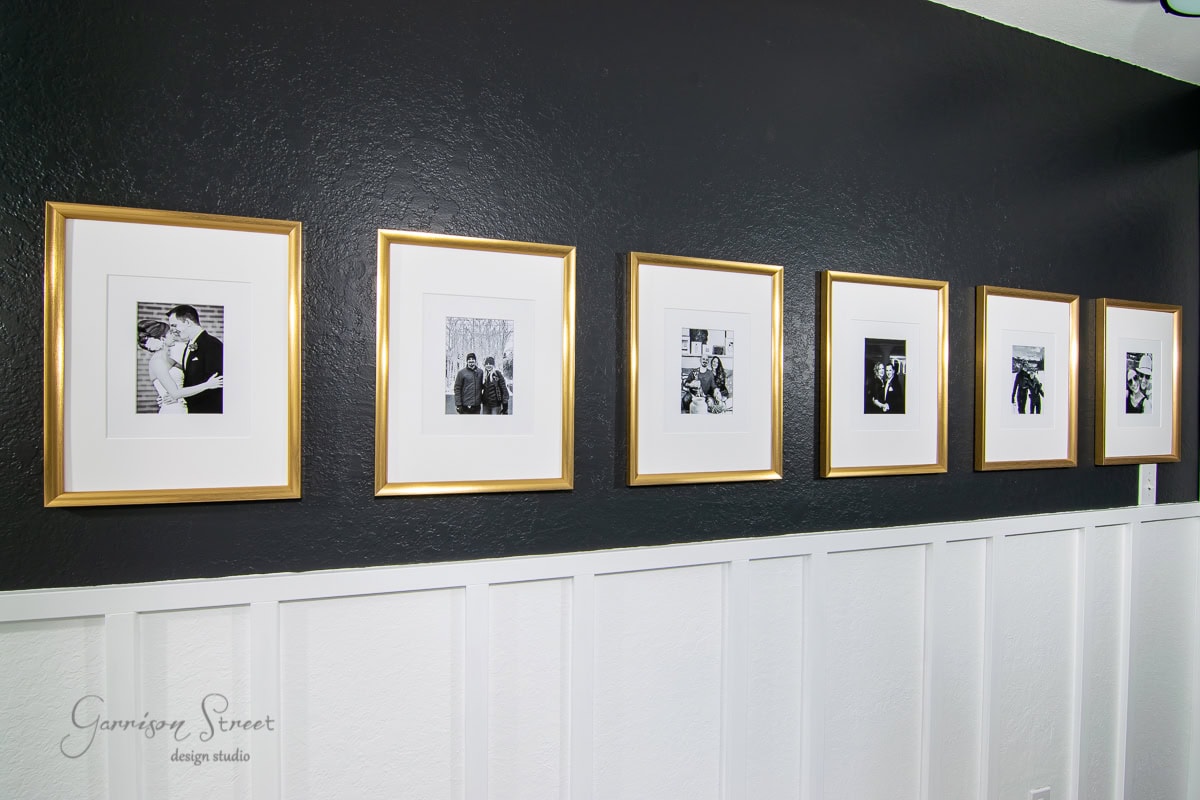 A series of six black and white photos in gold picture frames, accented with oversized mats, are neatly arranged on a black wall with white wainscoting. Each frame contains a different portrait, creating a cohesive, elegant display.