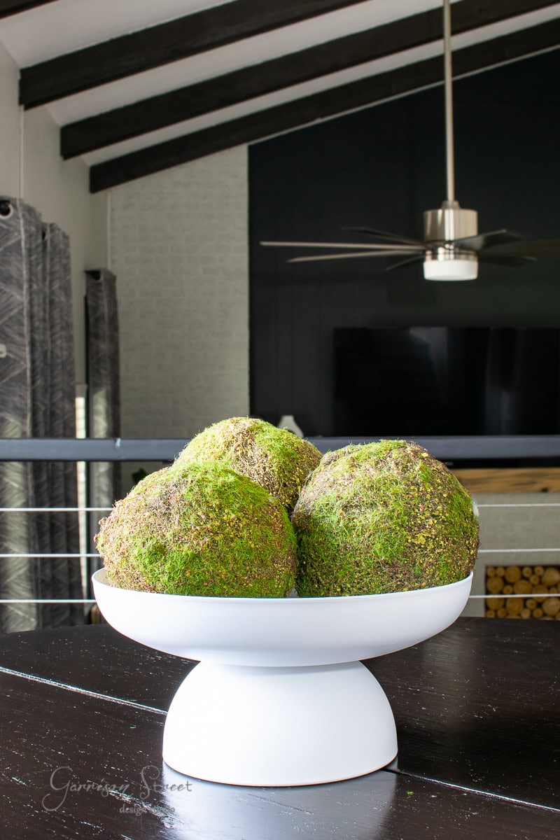 A white pedestal bowl on a dark wooden table holds three large decorative moss balls. The background features a modern interior with a ceiling fan and exposed beams, creating a stylish DIY aesthetic.