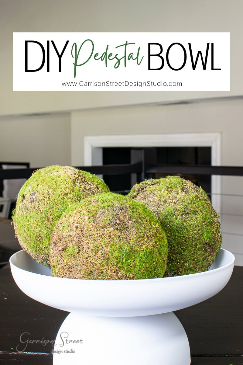A white pedestal bowl elegantly cradles three spherical, moss-covered objects, set against a backdrop of a partially visible room. At the top, "DIY Pedestal Bowl" is prominently displayed, accompanied by a website link below for further inspiration.