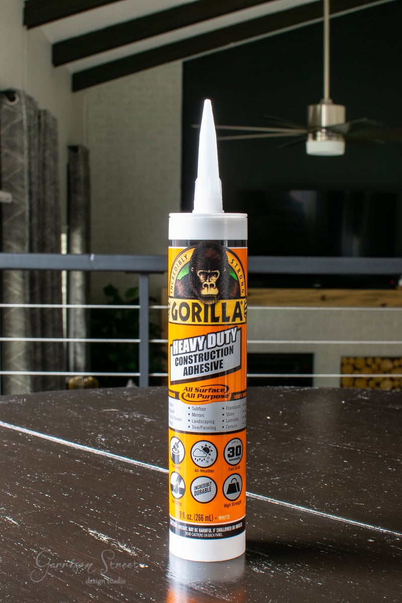 A tube of Gorilla Heavy Duty Construction Adhesive stands upright on a wooden surface, flanked by a stylish pedestal bowl. The label, showcasing a gorilla logo, is set against the backdrop of a modern living room equipped for any DIY project, complete with a TV and ceiling fan.