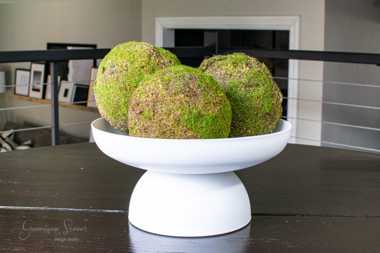 A white pedestal bowl on a dark wooden table holds three spherical moss-covered decorations. The background features a modern interior with framed art and soft lighting, creating a DIY atmosphere that invites creativity and elegance.