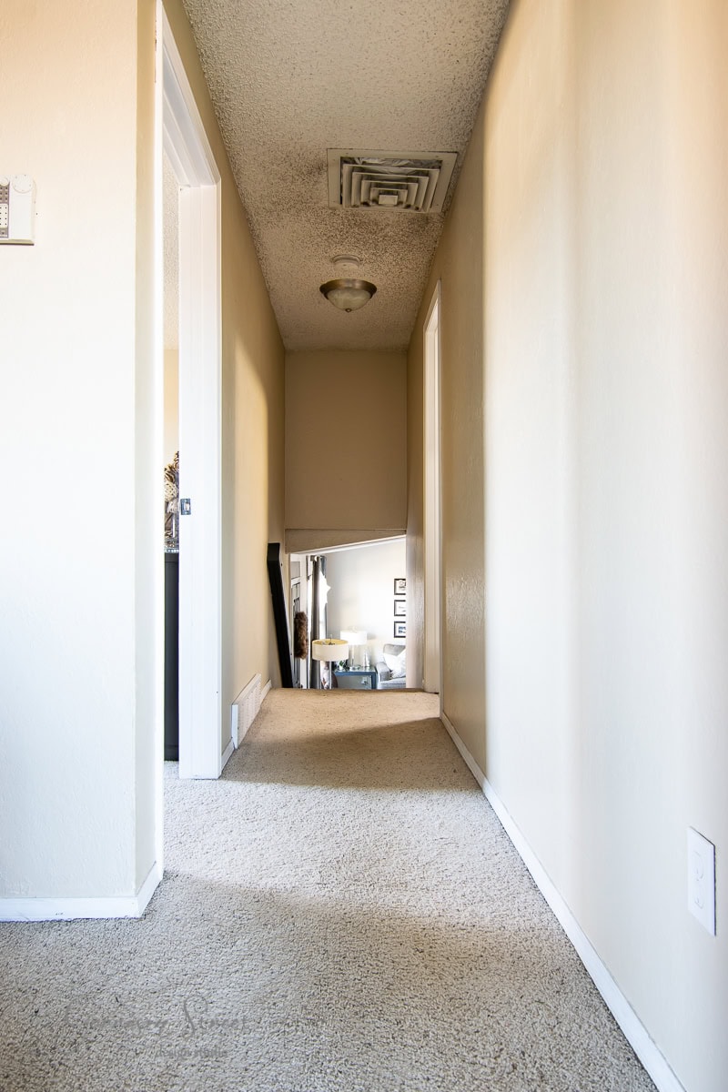 A long, narrow hallway adorned with board and batten walls features beige tones and carpet. The ceiling has a light fixture and vent. At the end of the hallway, a staircase leads down to a well-lit area with furniture partially visible. Doors are on the left and right.