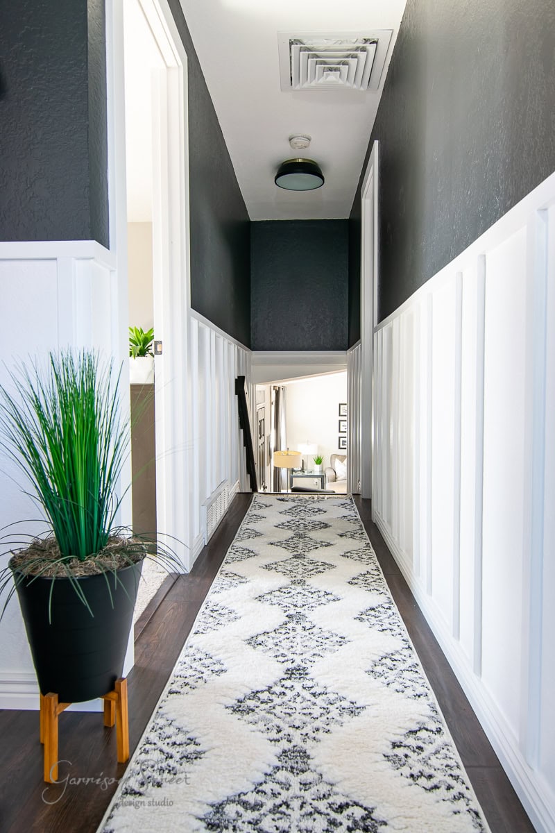 A modern hallway with dark walls and white board and batten wainscoting features a long, patterned rug leading to a bright room. A tall plant in a black pot stands on the left. Natural light illuminates the space from a doorway at the end.