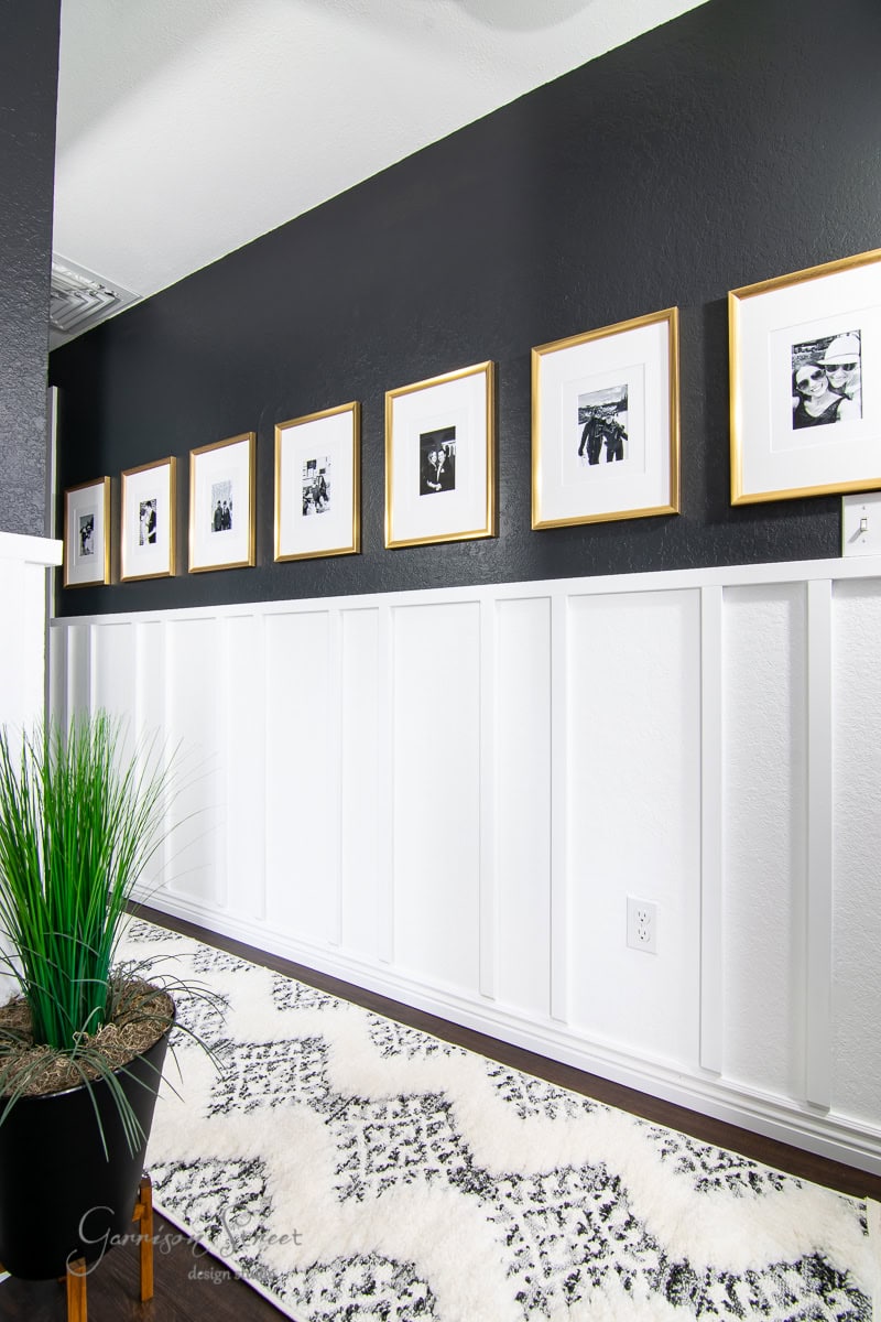 A stylish hallway features a black upper wall with white board and batten paneling below, adorned with eight evenly spaced gold-framed black-and-white photos. The floor has a white and black patterned rug, and a tall green plant sits in a brown pot.