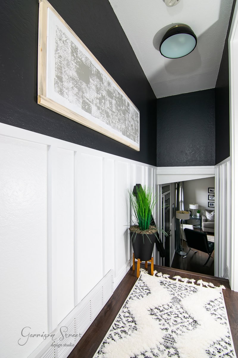 A modern hallway features a black and white color scheme with board and batten wall DIY detailing. White paneling contrasts with black upper walls, while a thin framed artwork hangs above. A plant in a black pot sits at the end, overlooking a dining area with a table and chairs.