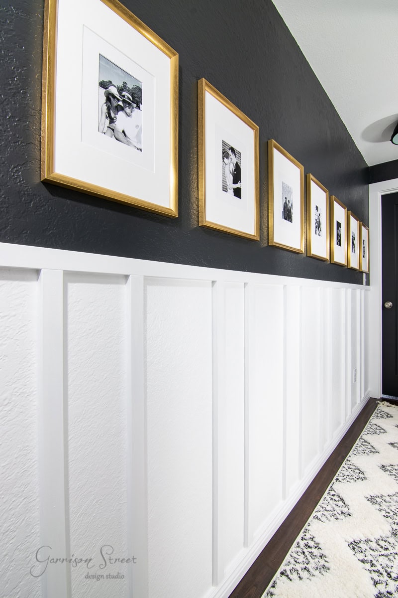 A hallway showcases a black and white wall adorned with framed black-and-white photos in gold frames. The upper wall is black, while the board and batten DIY lower section has white wainscoting. The wooden floor is complemented by a striking white and black patterned rug.