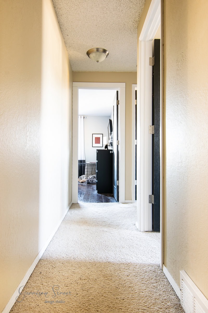 A narrow hallway with beige carpet and walls leads to a bright room with dark wood flooring. Several open doors line the hallway, and a black cabinet and framed picture are visible in the distant room.