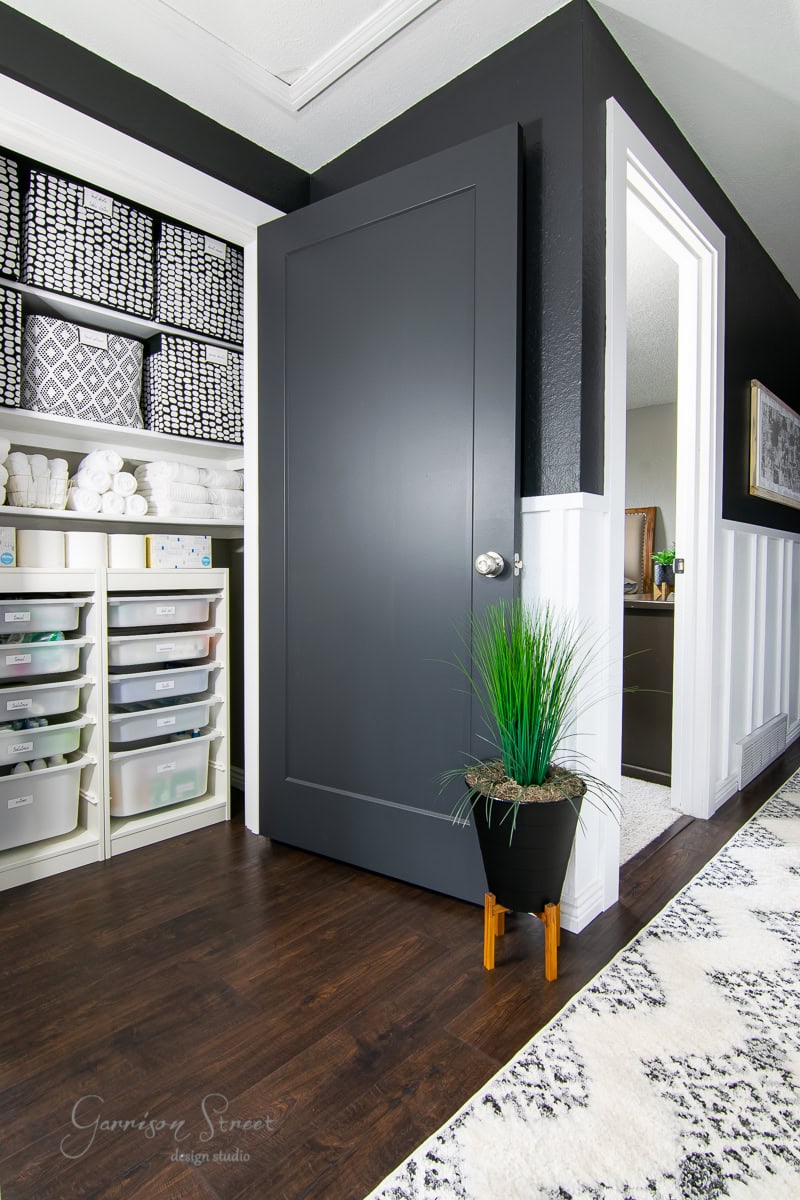 A modern storage room features a sleek black door and crisp white walls, enhanced by a stylish board and batten wall DIY. Shelves hold baskets and organized storage boxes while a decorative green plant sits on the dark wooden floor near the patterned rug.
