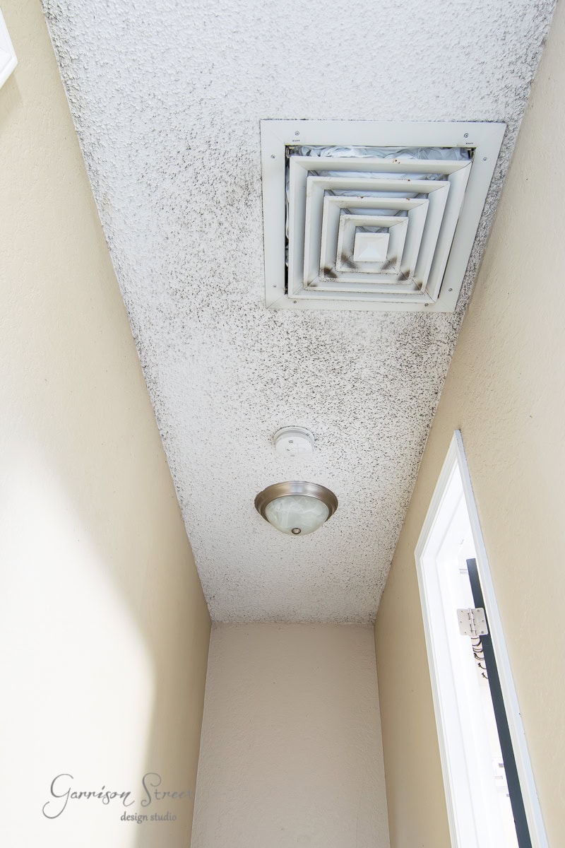 A hallway ceiling reveals patches of black mold around the light fixture and air vent, while beige walls loom, offering a stark contrast. A small window filters in natural light.